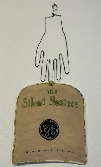 textile work featuring brown rectangle that says 'the silent hostess' with a wire hand above it